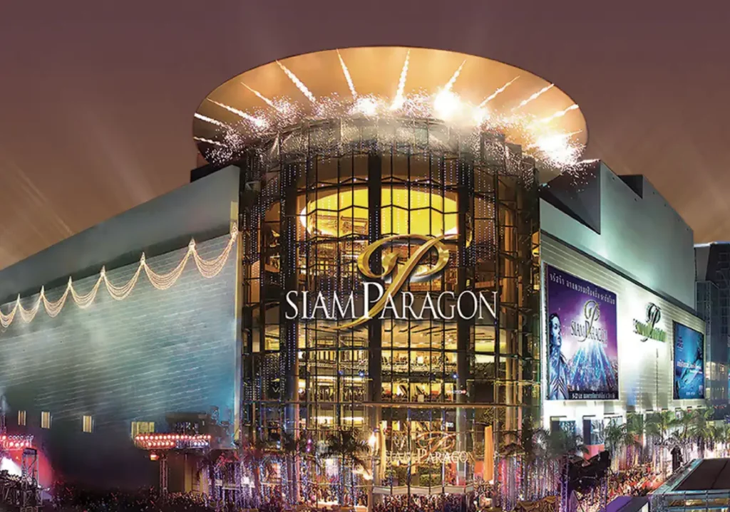 Escape the rain in Bangkok malls: Siam Paragon shopping center in Bangkok at night, featuring its distinctive curved glass facade and golden lighting