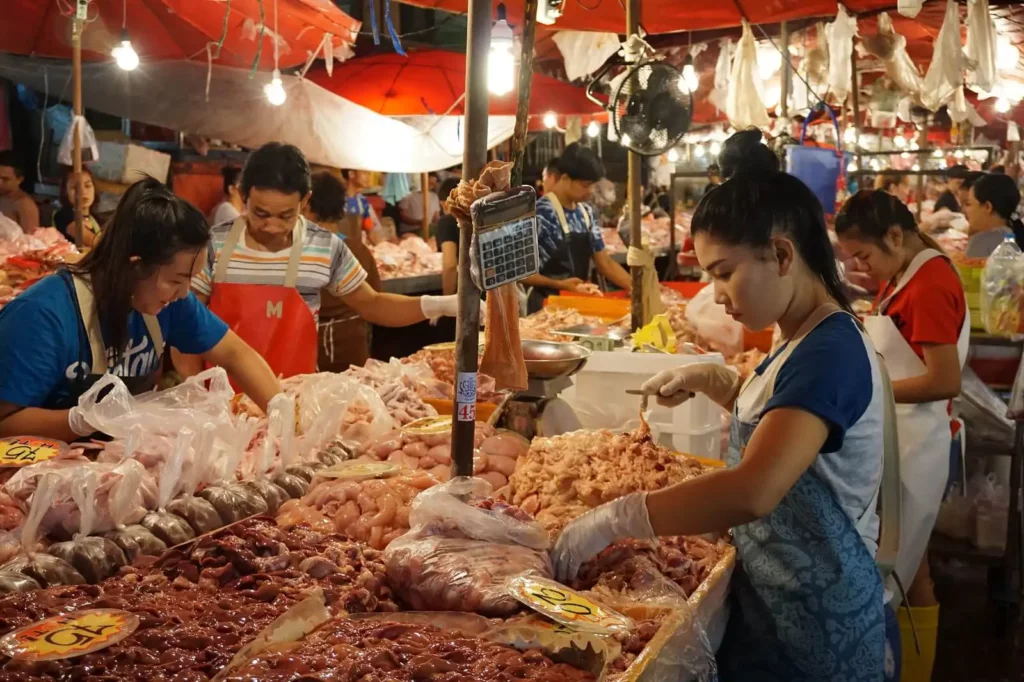 Bangkok Flower Markets: Fresh meat section at Khlong Toei Market with vendors in aprons preparing and selling various cuts of meat to customers under bright lighting