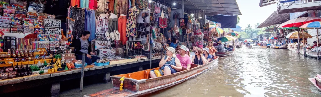 Top 10 Must-Do Activities in Bangkok: A group of tourists sits in a wooden boat while shopping at waterside market stalls selling textiles, souvenirs, and local goods at Damnoen Saduak floating market