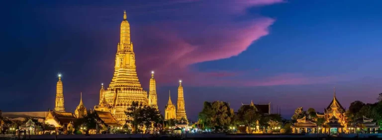 Top Photo Spots Bangkok: Spectacular evening shot of Wat Arun temple complex with its towering central spire and smaller prangs glowing golden against a dramatic purple and pink sunset sky over Bangkok