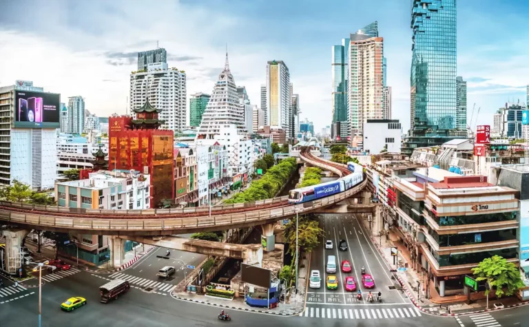 Smart Budget Travel Bangkok: Bangkok cityscape with the BTS Skytrain winding between modern high-rises and older buildings, while colorful taxis navigate the busy streets