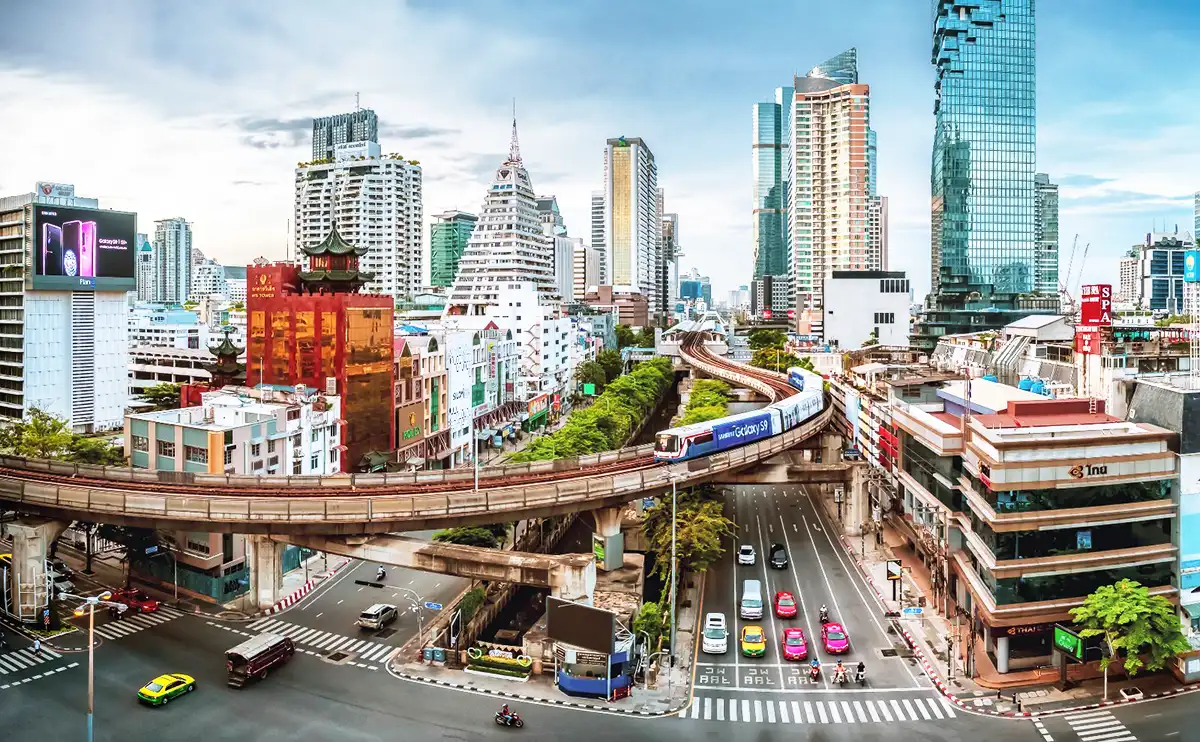 Smart Budget Travel Bangkok: Bangkok cityscape with the BTS Skytrain winding between modern high-rises and older buildings, while colorful taxis navigate the busy streets