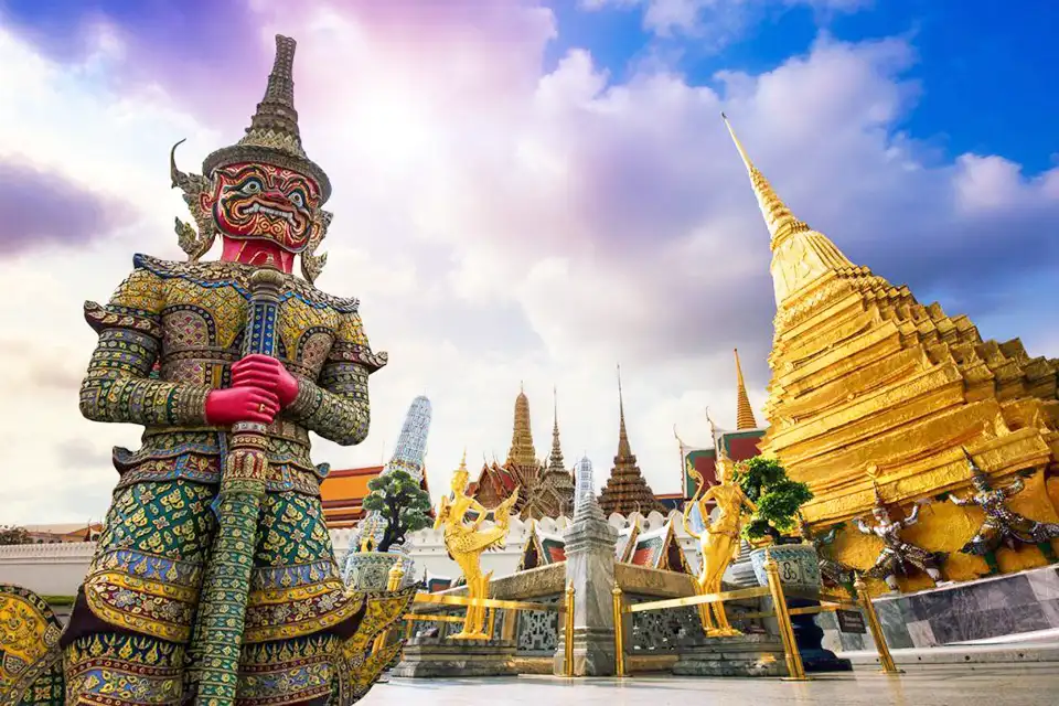 Bangkok FAQs: A towering Yaksha guards Wat Phra Kaew, with golden stupas and ornate temple spires glowing under a dramatic sky.