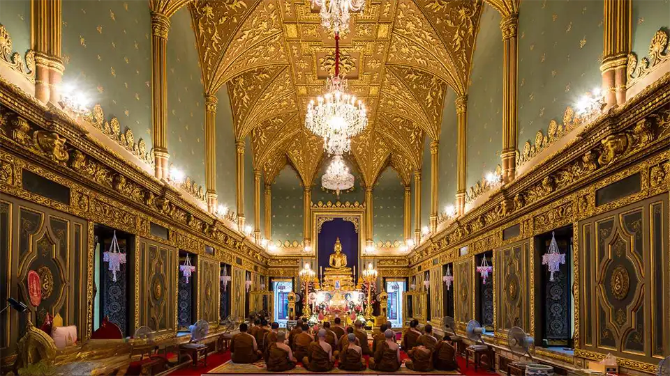 Grand interior of Wat Ratchabophit Sathit Maha Simaram with elaborate gold leaf ceiling, crystal chandeliers, and monks seated during a Buddhist ceremony