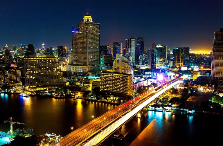 Modern Bangkok illuminated at night, with high-rises, fast traffic, and shimmering water.