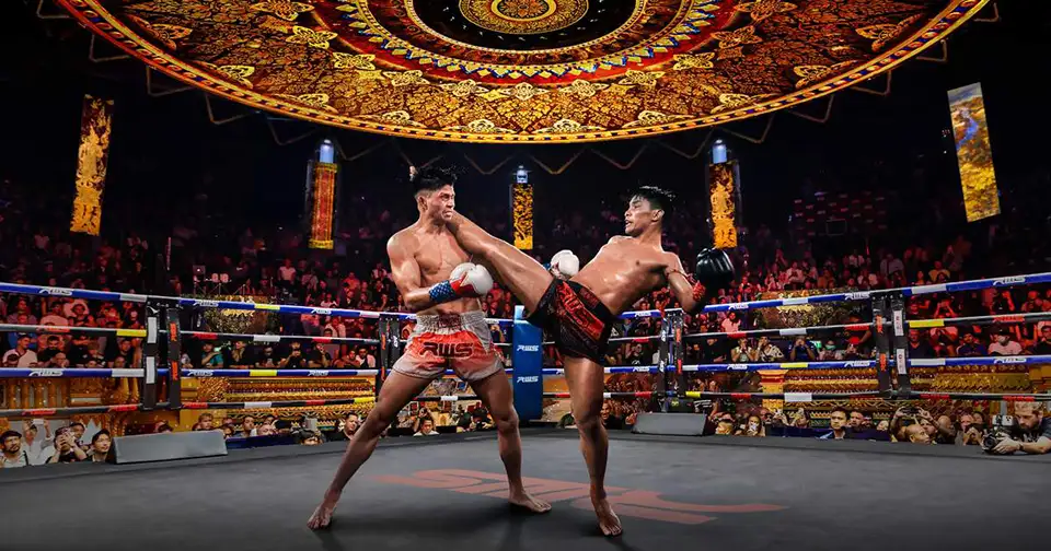 Two Muay Thai fighters clash in the ring, surrounded by dramatic architecture and cheering fans.