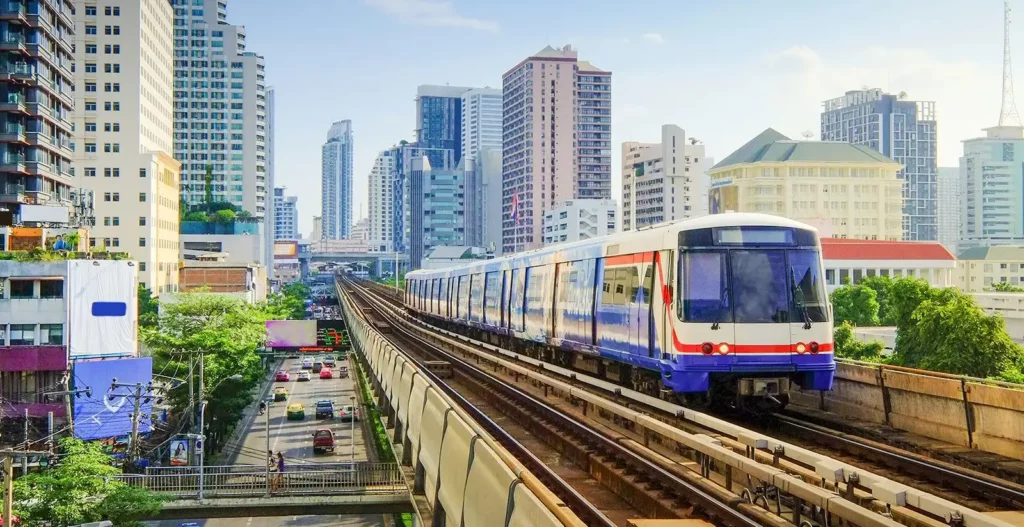 A Bangkok BTS train glides above city traffic, framed by high-rises and clear skies on a bright urban afternoon.