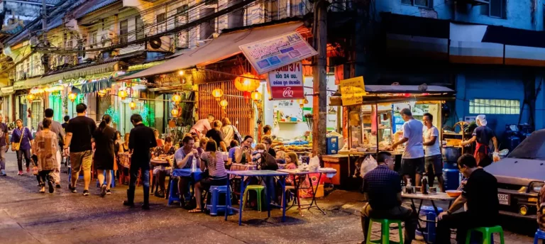 Top Bangkok Street Food: People enjoying food at a bustling Bangkok street food stall in the evening, with colorful lights and traditional shophouses in the background