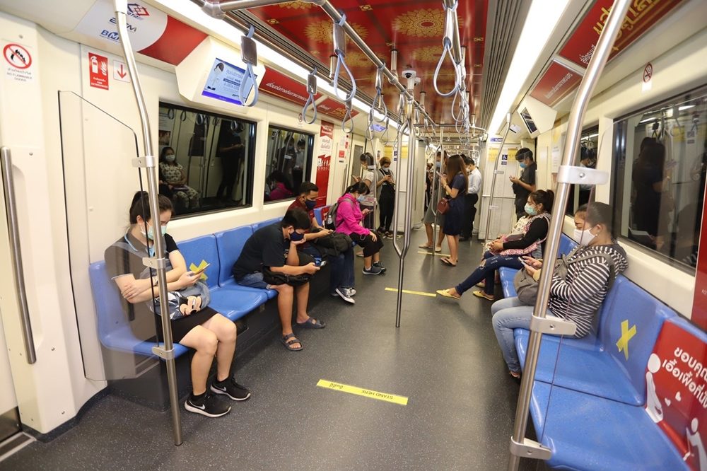 Inside an air-conditioned MRT subway car with passengers sitting on blue seats, some wearing masks, with yellow floor markers for social distancing