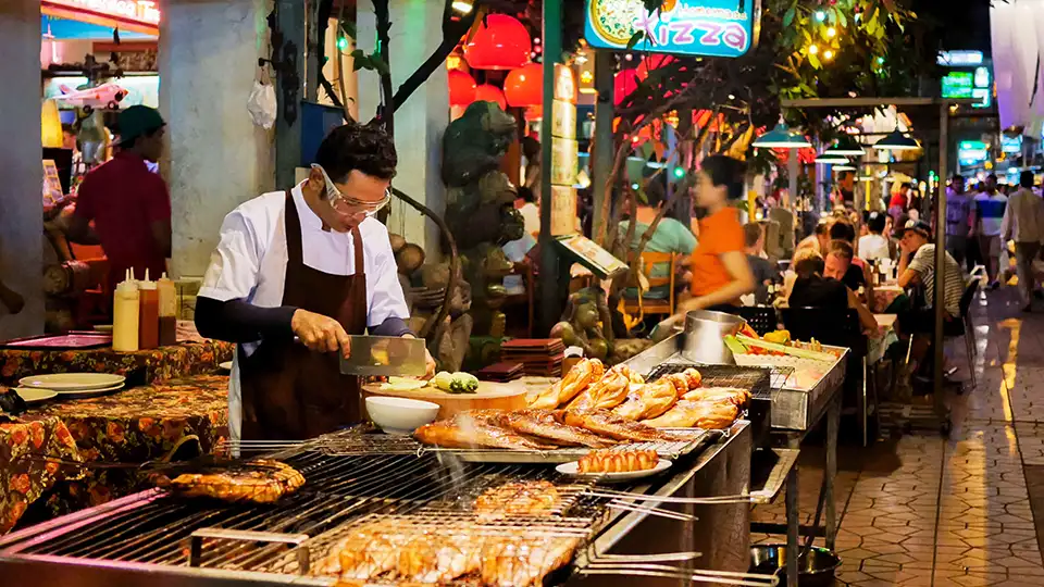 Bangkok FAQs: A street food vendor grills meats at a lively night market, surrounded by neon lights, red lanterns, and hungry diners.
