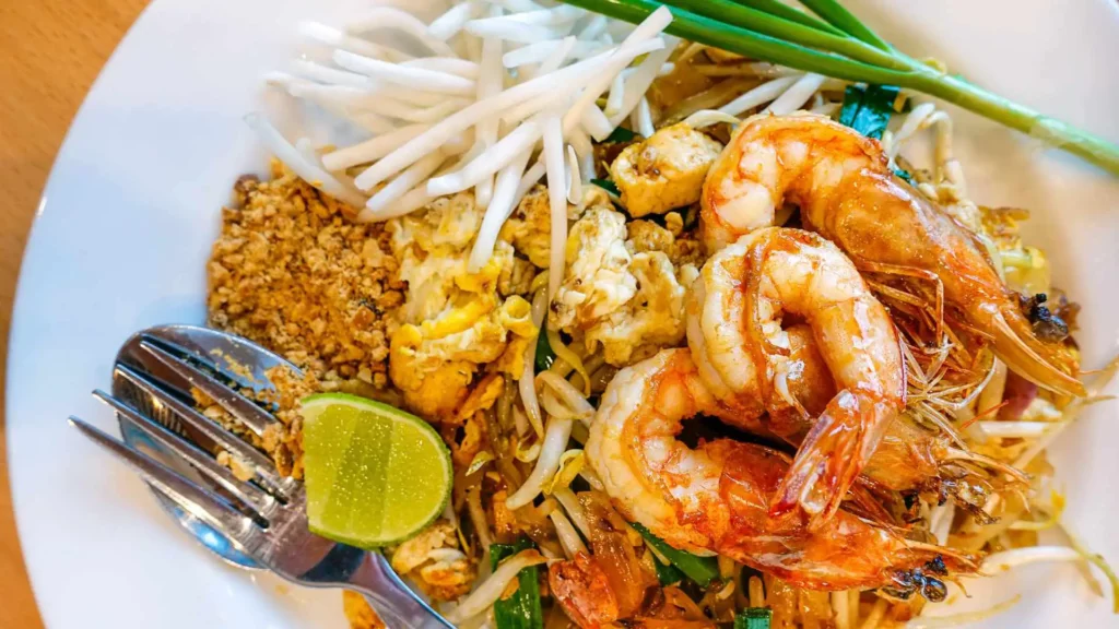 Bangkok FAQs: A plate of Pad Thai with shrimp, peanuts, and lime, ready to enjoy with a fork and spoon.