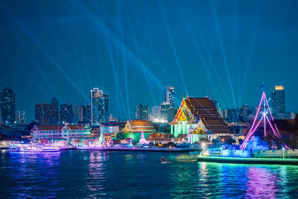 Night scene of traditional Thai temples lit in vibrant colors with illuminated royal barges on the Chao Phraya River, light beams in the sky, and modern Bangkok buildings in the distance during Vijit Chao Phraya 2025 celebration