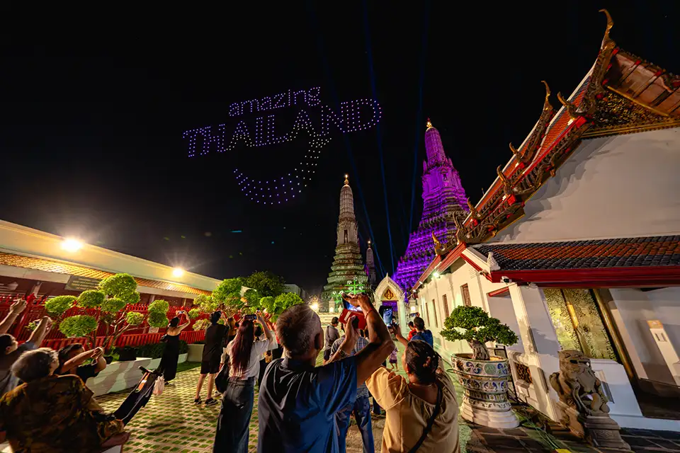 Crowd of visitors photographing a drone light display that reads 'Amazing Thailand' above the illuminated purple prang of Wat Arun temple during Vijit Chao Phraya 2025 festival