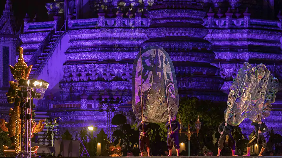 Performers holding large decorative Thai shadow puppets on stage with Wat Arun temple illuminated in purple light as the backdrop during Vijit Chao Phraya 2025 festival