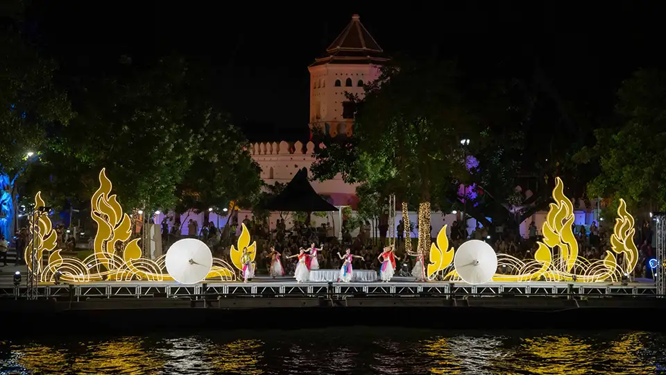 Dancers in traditional Thai costumes performing on an illuminated floating stage with golden flame decorations, with a historic fort tower in the background during Vijit Chao Phraya 2025 festival