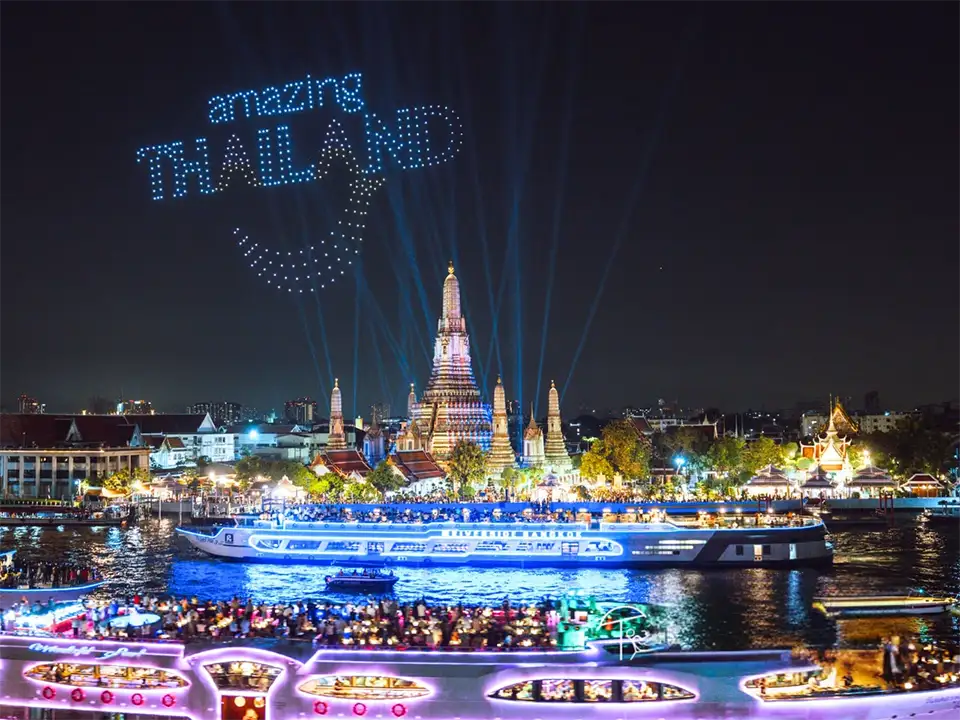 Spectacular night scene of the Chao Phraya River filled with brightly lit boats in blue, pink, and gold, with Wat Arun temple illuminated in the background, light beams shooting into the sky, and drones forming 'Amazing Thailand' text above during the Vijit Chao Phraya 2025 celebration