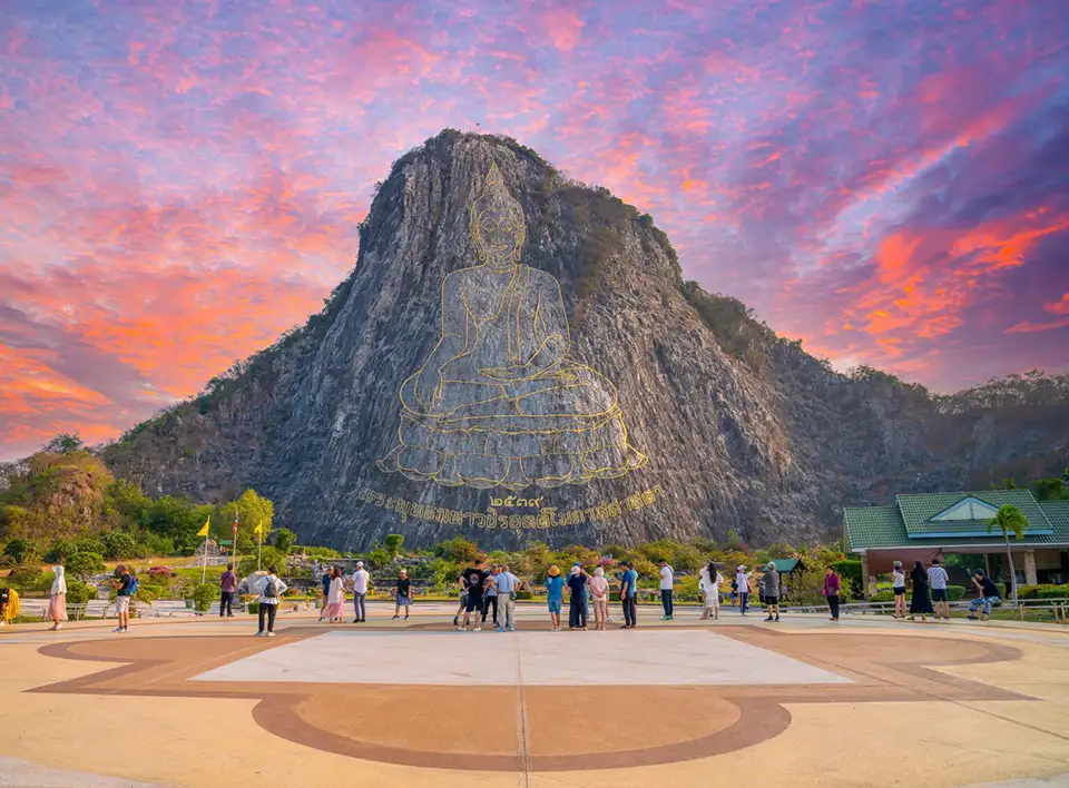 Tourists admiring the massive golden Buddha image carved into Khao Chi Chan mountain during a spectacular sunset in Pattaya