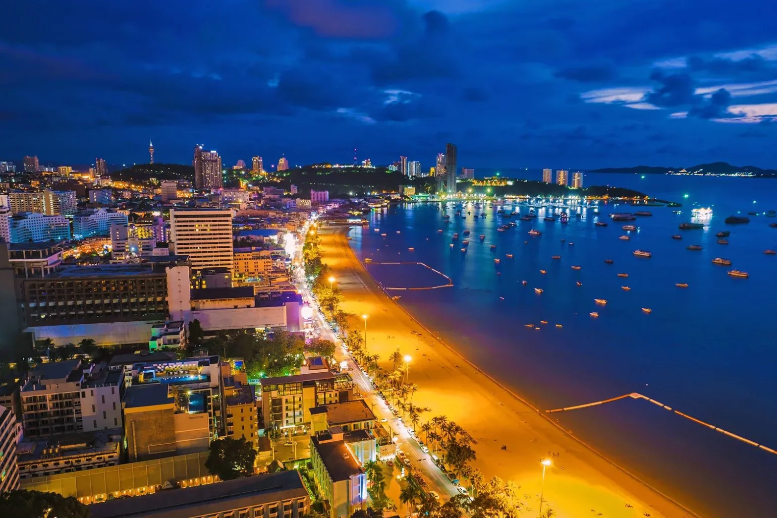 Aerial night view of Pattaya Beach, Thailand, with city lights, high-rise buildings, and boats along the coastline.