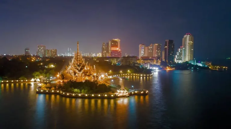 Night view of the Sanctuary of Truth temple in Pattaya, Thailand, illuminated against the city skyline with reflections shimmering on the water