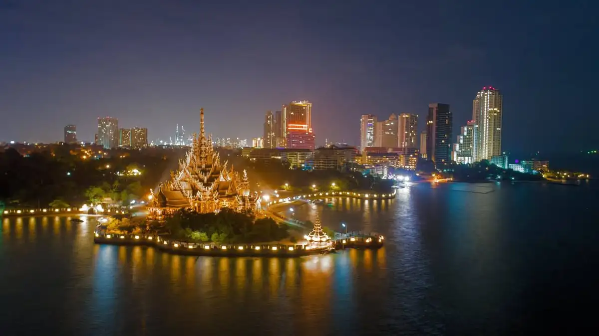 Night view of the Sanctuary of Truth temple in Pattaya, Thailand, illuminated against the city skyline with reflections shimmering on the water