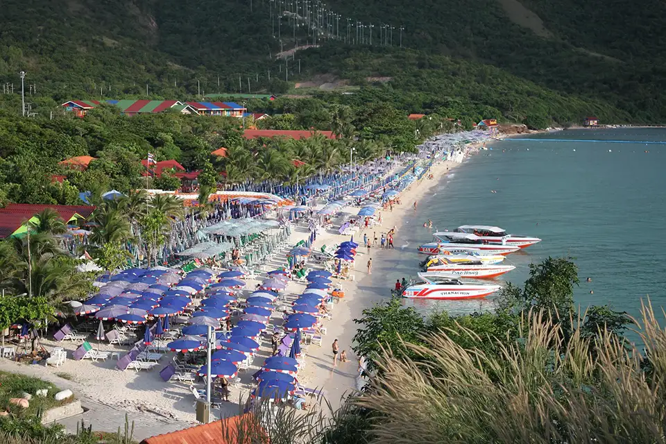 Popular Pattaya beach with rows of blue umbrellas, beachgoers, and speedboats docked along the shore
