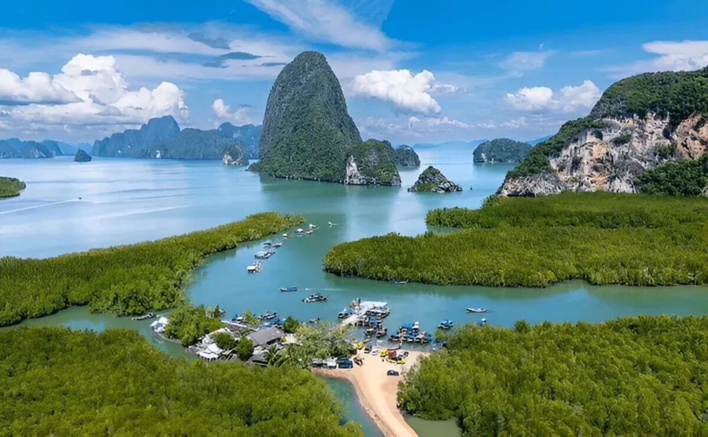 Aerial view of Phang Nga Bay showing limestone karsts rising from turquoise waters, with mangrove forests and traditional longtail boats anchored in a peaceful lagoon