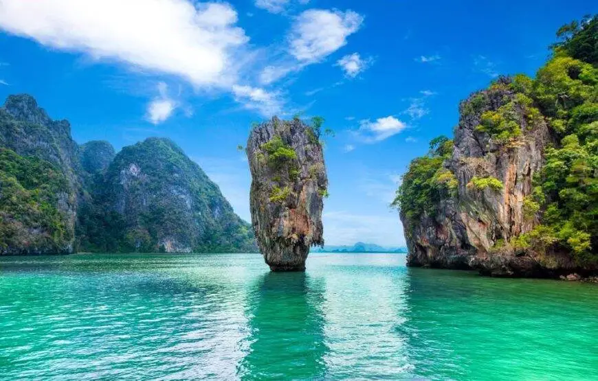 Crystal clear emerald waters surround towering limestone cliffs in Phang Nga Bay, with lush tropical vegetation clinging to the dramatic rock formations under a bright blue sky