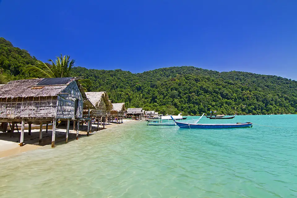 Traditional stilt houses and colorful longtail boats anchored in shallow turquoise waters along the pristine coastline of Phang Nga, with jungle-covered hills in the background