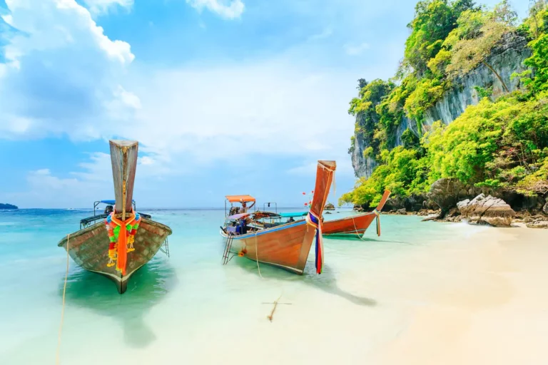Phuket Beach Two traditional Thai longtail boats with colorful ribbons moored in crystal-clear turquoise water at a pristine tropical beach, with dramatic limestone cliffs covered in lush green vegetation rising from the sea under a bright blue sky with white clouds