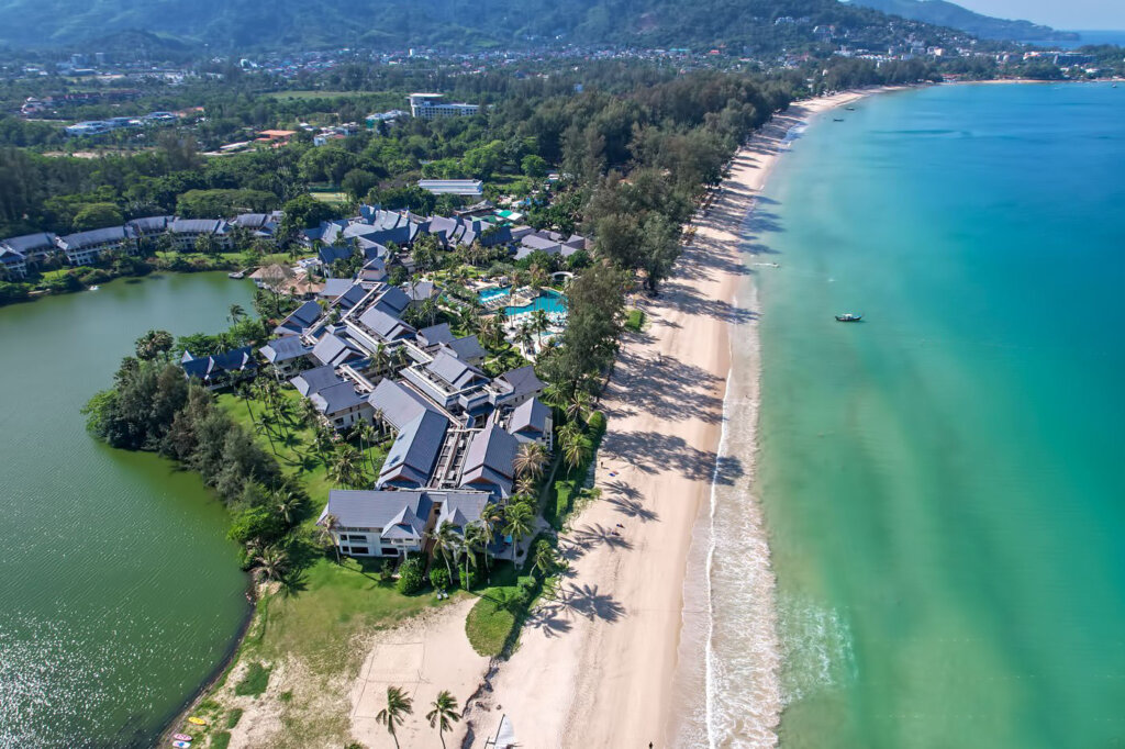 Phuket Bang Tao Beach Aerial view of a beachfront resort with swimming pools next to turquoise water and mountains in the distance.