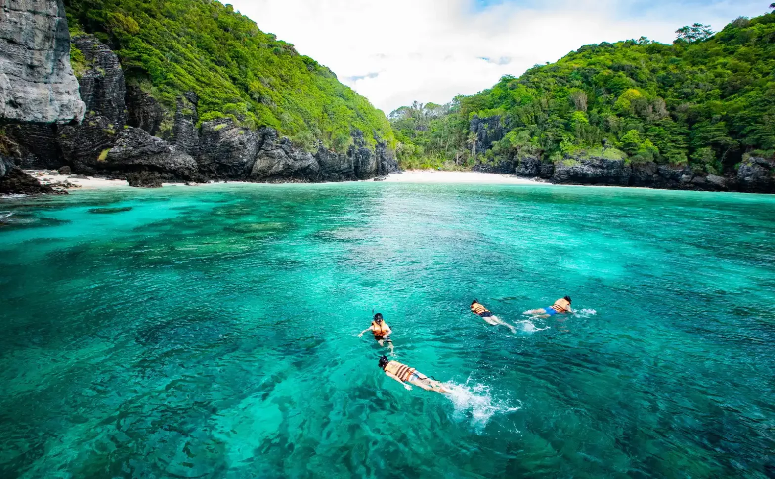Four people snorkeling in crystal-clear turquoise waters surrounded by lush green limestone cliffs and a secluded tropical beach