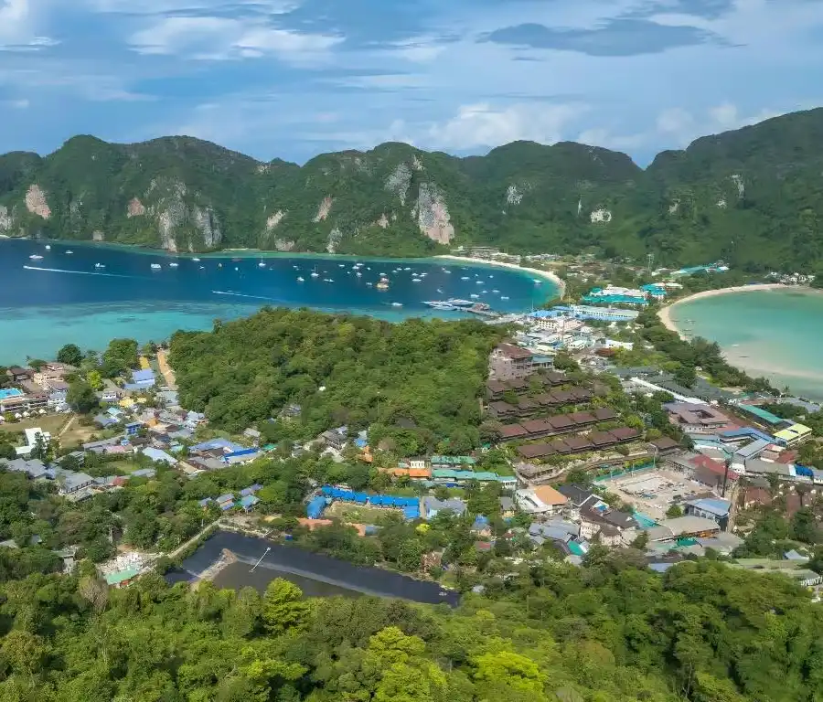 Phuket to Phi Phi Island Aerial view of a tropical bay with turquoise waters, white sand beaches, boats anchored offshore, and a small coastal town nestled between dramatic limestone mountains