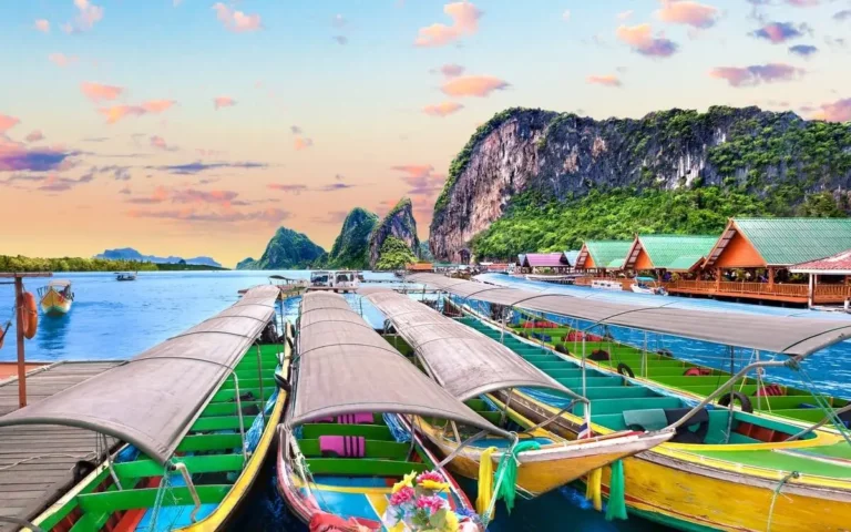 Colorful traditional longtail boats moored at a wooden pier in Phuket, with dramatic limestone karst mountains rising from turquoise waters in the background. The vibrant boats in green, yellow, and blue create a striking contrast against the peaceful bay, while floating restaurants and scenic cliffs complete this iconic Thai seascape under a pastel sunset sky.