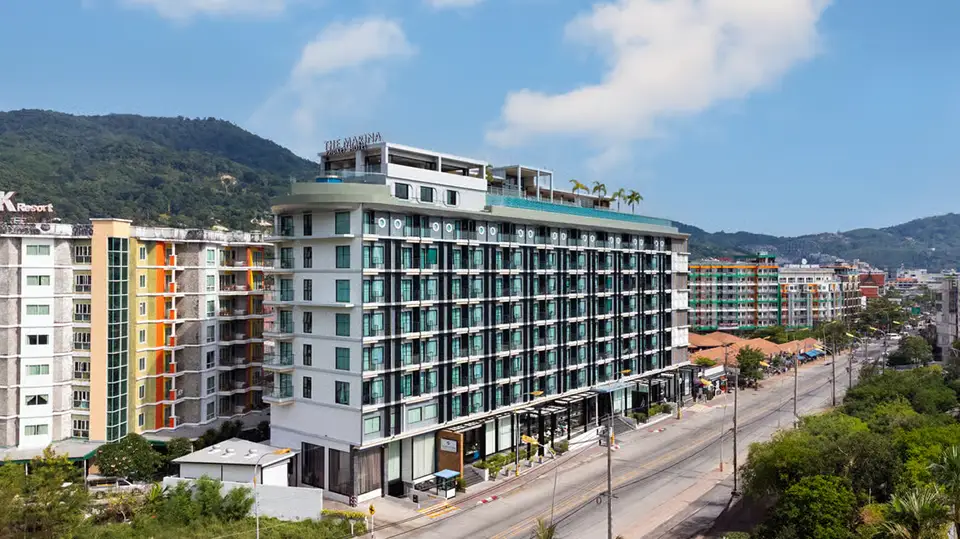 A modern multi-story hotel building in Phuket featuring contemporary architecture with glass balconies and clean white facades. The hotel sits prominently along a main road with lush green hills in the background, surrounded by other colorful buildings and tropical vegetation. The rooftop area appears to have additional amenities, while the ground level shows commercial spaces, representing the convenient urban location that makes this an ideal base for exploring Phuket.