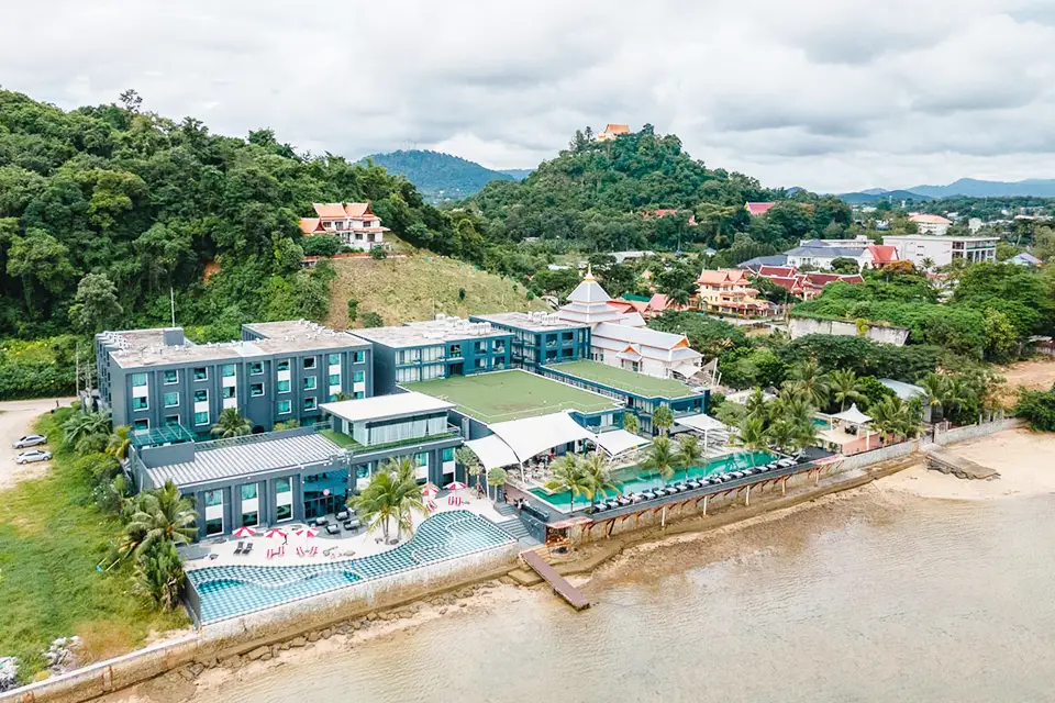 An aerial view of a modern beachfront hotel in Phuket featuring distinctive turquoise and white buildings arranged around a central courtyard with swimming pools. The contemporary resort sits directly on a sandy beach with calm waters, surrounded by lush tropical hills and local residential areas. Palm trees dot the property while the multi-story accommodation blocks offer ocean views, showcasing the perfect blend of modern amenities and natural tropical beauty.