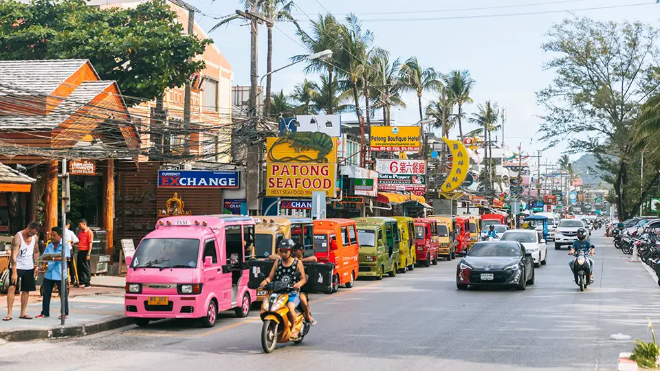 A bustling street scene in Phuket showing colorful food trucks and local vendors lined up along the main road. The vibrant pink, orange, and green mobile food stalls create a lively atmosphere while tourists and locals gather around to sample street food. Traditional Thai signage advertises local seafood restaurants like "Patong Seafood," and palm trees frame the scene against a partly cloudy sky. Motorcycles and cars navigate the busy street filled with the energy of local commerce and tourism.