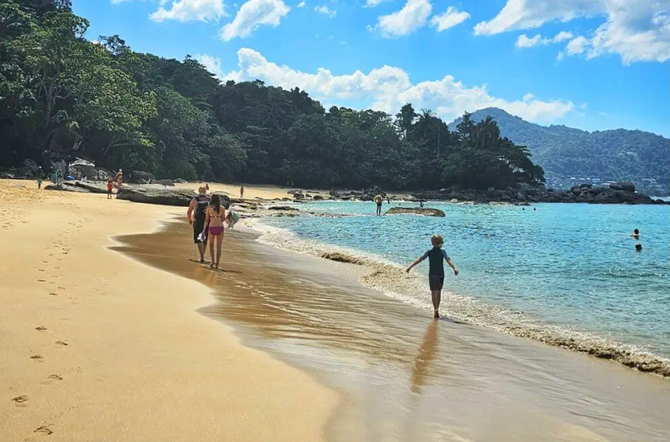 Hidden Beaches in Phuket: Tourists walking along the golden sand at Banana Beach, Phuket with turquoise water and lush hills in the background.