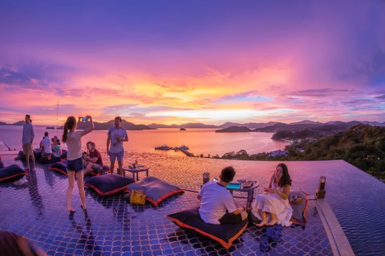 Guests enjoying sunset drinks at Phuket Top View Bar's infinity pool deck overlooking Cave Panwa, with people relaxing on floating bean bags and taking photos of the golden hour view