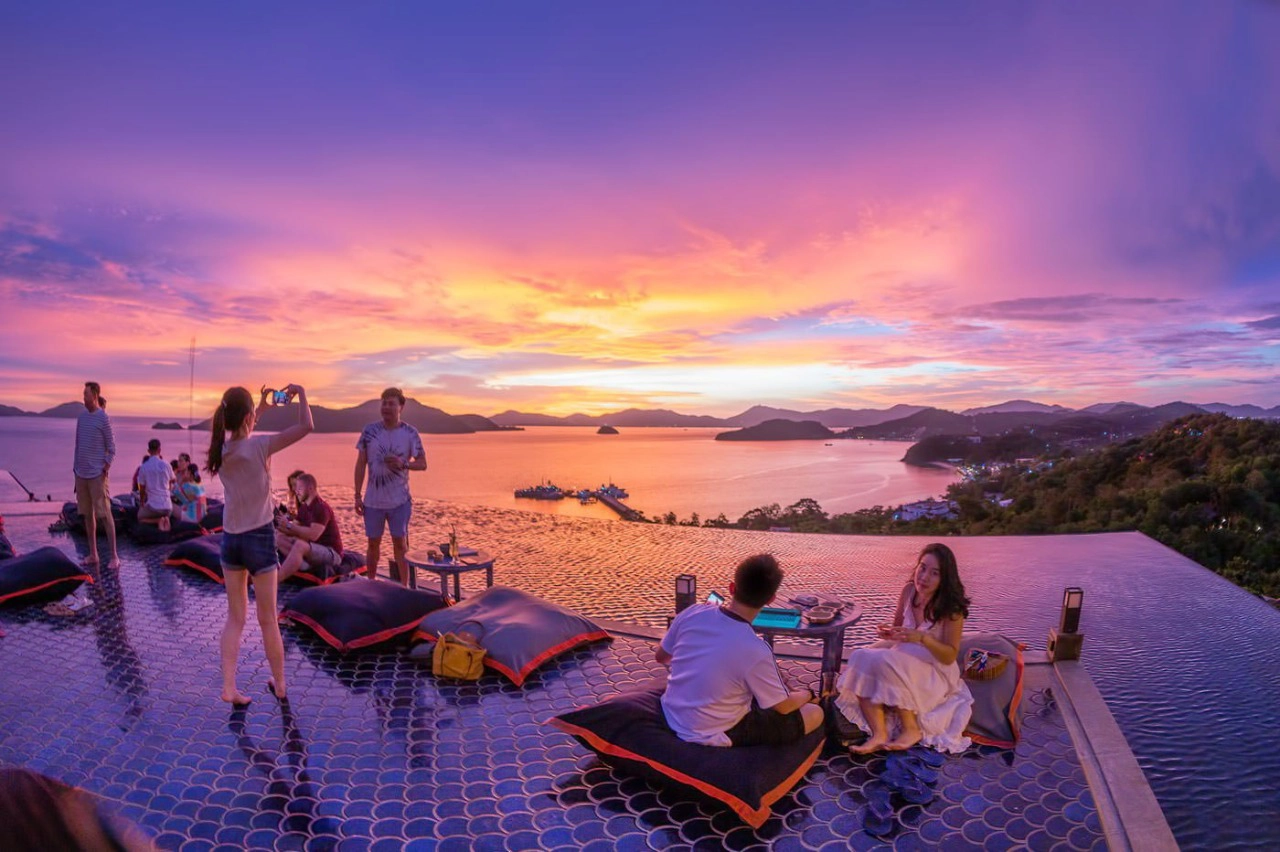Guests enjoying sunset drinks at Phuket Top View Bar's infinity pool deck overlooking Cave Panwa, with people relaxing on floating bean bags and taking photos of the golden hour view