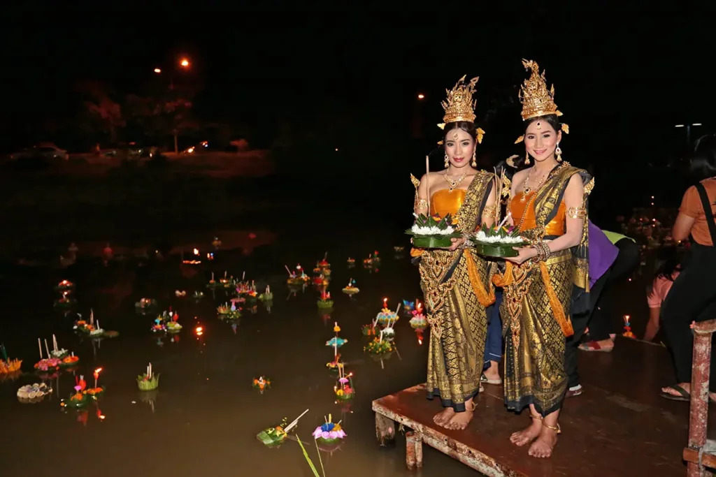 Women in ornate Thai costumes releasing krathongs into water during nighttime Loy Krathong festival