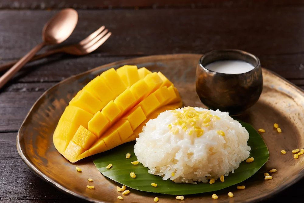 Thai food: Classic Thai dessert of sweet coconut sticky rice served with fresh mango slices and coconut milk, presented on a wooden plate with banana leaf