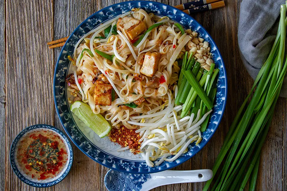 Thai food: Traditional Pad Thai served on a decorative blue plate with lime wedge, crushed peanuts, and fresh scallions on the side