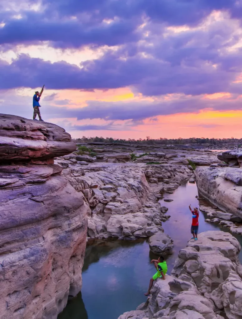 Hidden Thailand: Visitors exploring eroded rock formations under a dramatic sunset sky at Hat Chom Dao, Ubon Ratchathani