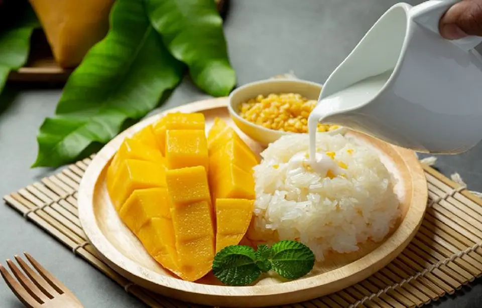 Thai Snacks: Traditional Thai mango sticky rice served with coconut drizzle, crispy mung beans, and fresh mint on a wooden plate.